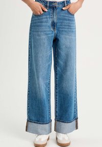 Jeans de mezclilla azul de pierna ancha con una cintura alta, con dobladillo doblado y costuras en contraste. Combinados con zapatillas blancas.