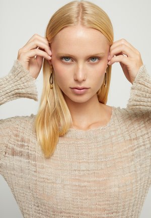 Mujer rubia con ojos azules ajusta pendientes colgantes dorados, vistiendo un suéter beige de punto suelto contra un fondo claro y liso.