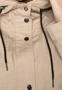 Beige Jacke mit Kapuze, sicherer Toggle-Verschluss, schwarzen Kordeln, glänzenden schwarzen Knöpfen und einer glatten, leichten Stofftextur.