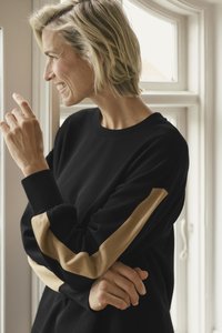 Schwarzer Sweatshirt mit kontrastierenden beigen Streifen an den Ärmeln. Rundhalsausschnitt, weicher Stoff und lockere Passform. Natürliches Licht von einem Fenster.