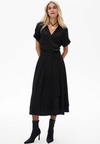 Schwarzes Wickelkleid aus glattem Stoff, kurze Ärmel, taillierter Schnitt, knielanger Rock, kombiniert mit schwarzen Stiefeln mit Absatz und einer dekorativen Halskette.