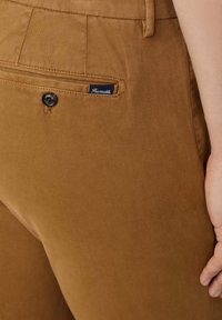 Pantalones de algodón marrones que cuentan con un bolsillo trasero con un botón negro y una etiqueta de la marca "Faconnable". La tela tiene un acabado texturizado.