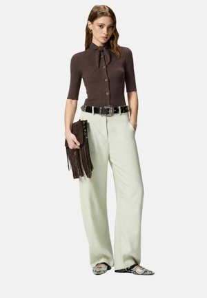 Donna che indossa un top marrone a maniche corte abbottonato con cravattino al collo, pantaloni chiari a gamba larga, cintura nera, pochette con frange e scarpe appuntite in pelle di serpente.