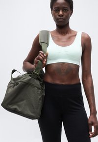 Αθλητικός άνθρωπος που φοράει ελαφρύ αθλητικό μπουστάκι και μαύρο κολάν, κρατώντας πράσινη τσάντα γυμναστηρίου στον ώμο, δείχνοντας τατουάζ λιονταριού στον κορμό.