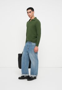 Zöld texturált, hosszú ujjú póló, világoskék bővített farmernadrág felgöngyölt szárakkal, fekete bőr cipő, és egy fekete táska.