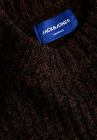 Étiquette en tissu bleu avec texte blanc « Jack & Jones Originals » cousue sur un vêtement en laine tricotée marron foncé.