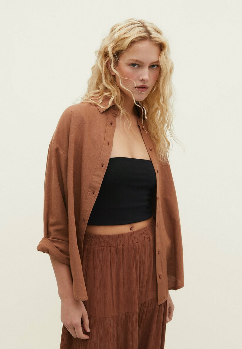 Chemise marron à boutons en tissu léger, portée par-dessus un crop top noir, associée à une longue jupe brune texturée. Cheveux naturels et ondulés.