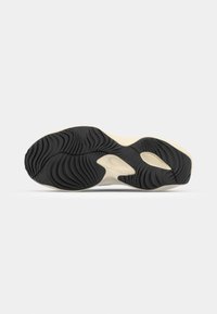 Semelle d'une basket beige et noire vue par en dessous avec des motifs courbés et rainurés sur fond blanc.