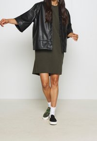 Czarna, sztuczna skórzana, oversize'owa koszula, oliwkowa sukienka, białe skarpetki do kostek oraz kamuflażowe czarne/zielone sneakersy. Prosty, nowoczesny design.