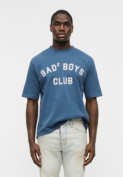 Άνδρας που φοράει μπλε μπλουζάκι με την επιγραφή "BAD² BOYS CLUB" και τζιν ανοιχτού χρώματος, στέκεται μπροστά σε απλό λευκό φόντο.