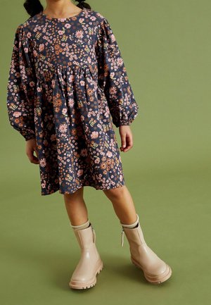 Robe à motifs floraux avec des manches bouffantes en bleu marine, ornée de fleurs roses, oranges et blanches. Associée à des bottes de cheville imperméables beiges.