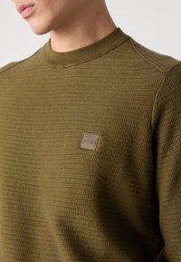 Sweter dresowy w kolorze oliwkowej zieleni z teksturowanym wykończeniem. Posiada okrągły dekolt oraz małą, kwadratową naszywkę z logo na piersi.