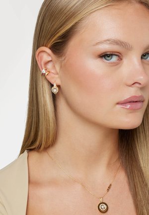 Boucles d'oreilles en or avec des pierres précieuses transparentes et un collier en chaîne délicate avec un pendentif circulaire sur un fond de teint naturel.