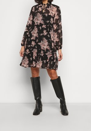 Femme portant une robe noire à fleurs à manches longues avec un col à nouer et des bottes noires jusqu'aux genoux, debout devant un fond clair et uni.