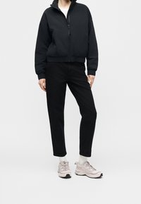 Personne portant une veste noire zippée, un pantalon noir, des chaussettes blanches et des chaussures de sport rose clair, debout sur un fond uni.