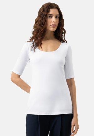 Donna con capelli castani ondulati che indossa una maglietta bianca a maniche corte con scollo rotondo e pantaloni scuri, in piedi davanti a uno sfondo chiaro uniforme.