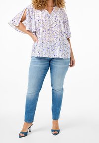 Blouse violet clair à imprimé floral, manches courtes et col à volants. Associée à un jean bleu ajusté et des escarpins bleus à bout ouvert.
