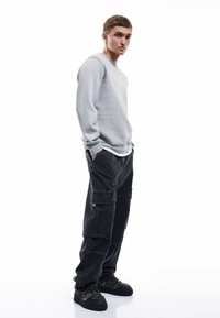 Sudadera gris con cuello redondo, combinada con pantalones cargo negros que tienen múltiples bolsillos y un corte holgado. Zapatillas negras con detalle texturizado.