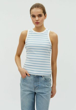 Mladá žena nosí tielkové svetlomodro-biele pruhované tričko a voľné svetlomodré rifle, s jednou rukou v vrecku.