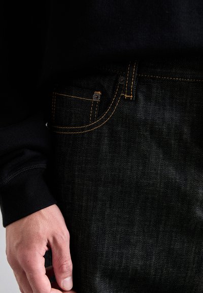 Mână sprijinită lângă buzunarul din față al unei perechi de blugi negri cu cusături portocalii, purtând un tricou negru cu mânecă lungă.
