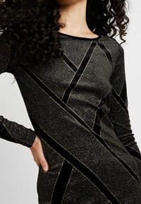 Svart långärmad klänning med ett geometriskt mönster av rhinestones och sammetstrim, med en figurnära silhuett och rundad halsringning.