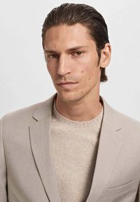 Light beige suit jacket over a light tan sweater, smooth fabric, structured design, and a narrow lapel with an accent pocket detail.