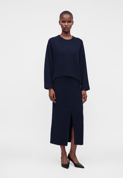 Femme debout portant un pull bleu marine à manches longues et une jupe midi assortie avec une fente à l'avant, associée à des talons aiguilles noirs pointus.