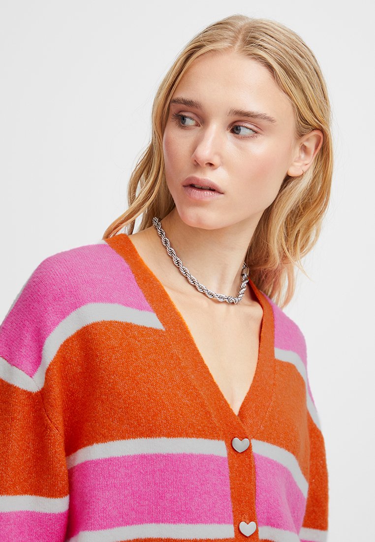 Młoda kobieta z blond włosami, nosząca pomarańczowo-różowy kardigan w paski z guzikami w kształcie serca oraz srebrny naszyjnik z skręconego łańcuszka.