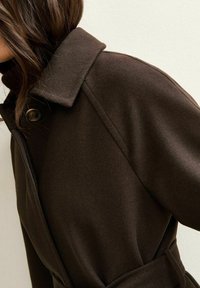 Bruine wollen jas met kraag, knoopdetails en riem. Soepele textuur, nauwsluitende pasvorm, getoond op een model tegen een neutrale achtergrond.