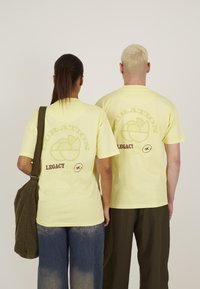 Tricouri din bumbac galben cu imprimeu grafic pe spate. Designul include "CURATION" și "LEGACY STU 2024" în maro. Croială lejeră, purtate cu blugi largi.