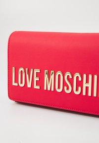 Rød kunstlær lommebok med gull "LOVE MOSCHINO" bokstaver, glatt tekstur og kontrasterende søm langs kantene.