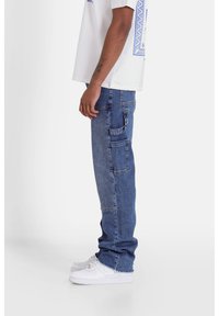 Pantalones cargo de denim azul claro con bolsillos laterales y diseño de pierna recta, combinados con zapatillas blancas y una camisa blanca de manga corta.