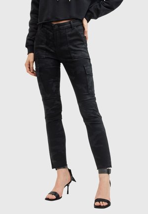 Frau trägt schwarze Cargohosen mit Camouflage-Muster und Knienähten sowie schwarze Sandalen mit hohen Absätzen, Hand auf der Hüfte ruhend.