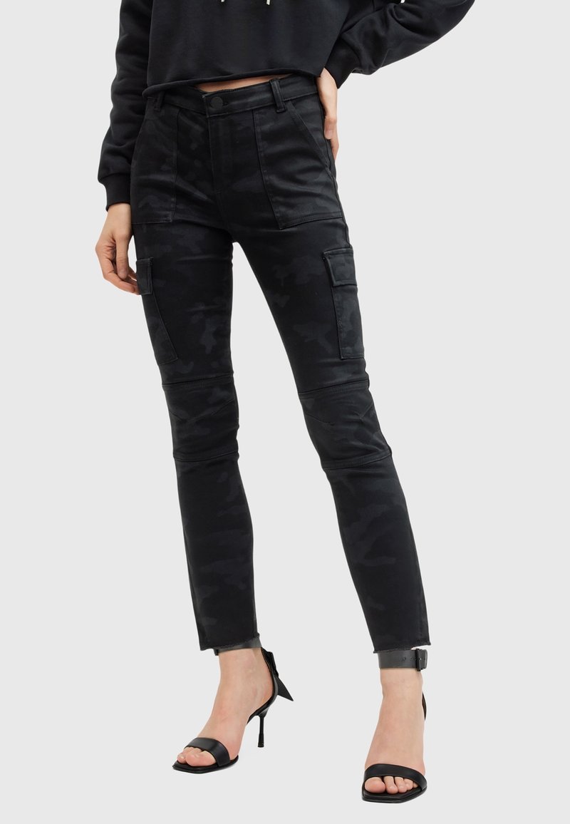 Femme portant un pantalon cargo camouflage noir avec des coutures aux genoux et des sandales à talons hauts noires, main posée sur la hanche.