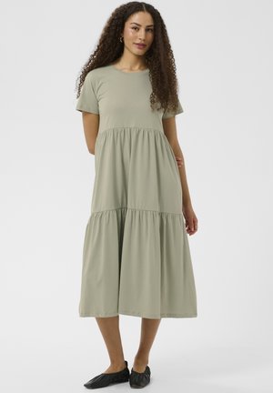 Moteris su garbanotais plaukais, dėvinti žalsvos žalios spalvos trumpomis rankovėmis sluoksniuotą midi suknelę ir juodus plokščius batelius, stovinti prie paprasto šviesaus fono.