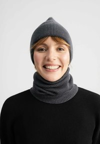 Graue gerippte Mütze mit spitzem Oberteil, kombiniert mit einem passenden gerippten Halswärmer aus einem gemütlichen Stoff, getragen über einem schwarzen Pullover.