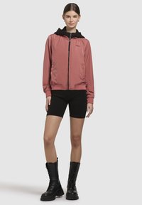 Veste bomber rose avec une capuche noire, fermeture éclair à l'avant et poches latérales. Associée à un short noir et des bottes montantes noires à lacets.