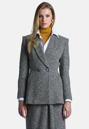 Femme aux longs cheveux blonds portant un blazer gris à double boutonnage, une jupe assortie, une chemise blanche, un col roulé moutarde et de grandes boucles d’oreilles en forme d’anneaux dorés.