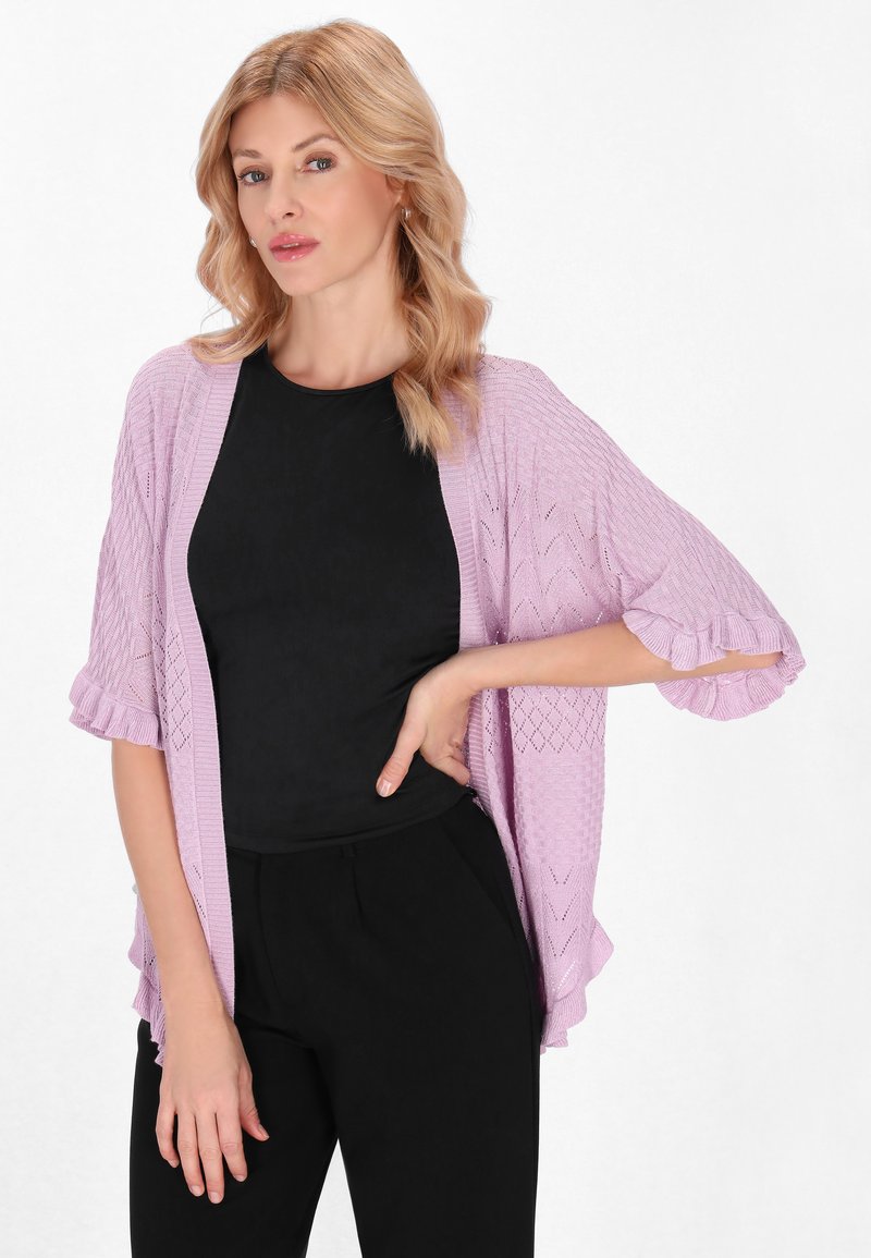 Cardigan lavorato a maglia di un leggero colore viola con un motivo testurizzato, con apertura frontale e maniche arricciate, indossato sopra un top aderente nero e pantaloni neri.