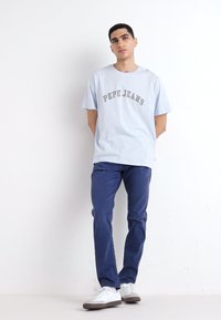 Svijetloplava pamučna majica s natpisom "PEPE JEANS" ispisanim sivo, uparena s tamnoplavim hlačama. Model nosi bijele tenisice. Jednostavna pozadina.
