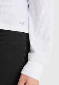 Vit långärmad tröja med ett strukturerat stickat mönster. Har ribbade ärmslut och en liten logotyp i silver nära fållen. Svart tyg under.