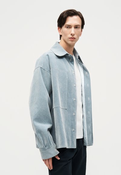 Νεαρός άνδρας που κοιτάζει δεξιά, φορώντας μπλε ανοιχτόχρωμο πουκάμισο με κουμπιά πάνω από λευκό T-shirt και μαύρο παντελόνι, μπροστά σε απλό φόντο.