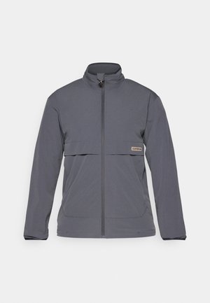 Veste zippée grise en tissu léger. Dispose d'un col haut, de poches frontales et d'une poche horizontale sur la poitrine avec une étiquette de logo.