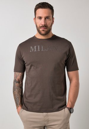 Mann mit Bart und tätowiertem linkem Arm, trägt ein braunes "MILAN"-T-Shirt und beige Hose, steht mit den Händen in den Taschen.