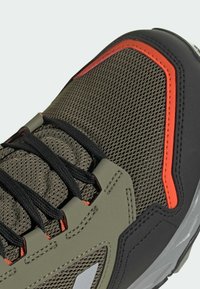 Gros plan d'une chaussure de sport avec des lacets noirs, un mesh vert olive, des surcouches noires et grises, et des détails orange.