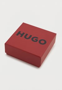 Κόκκινο ορθογώνιο κουτί με ματ φινίρισμα και τη διακριτική επιγραφή Hugo σε έντονα μαύρα γράμματα στην κορυφή. Συμπαγής σχεδιασμός, κατάλληλο για δώρο.