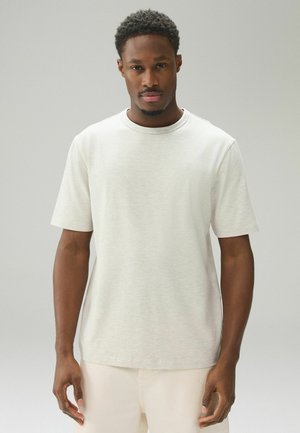 Mand med kort krøllet hår iført en ensfarvet lysebeige kortærmet t-shirt og lyse bukser, stående mod en neutral grå baggrund.