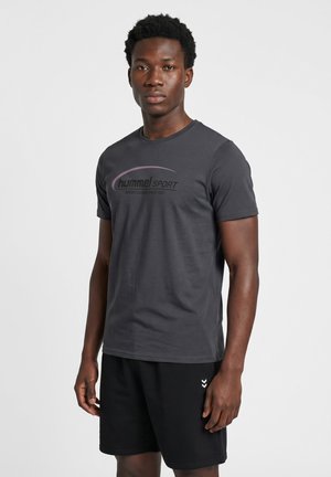 Junger Mann, der ein dunkelgraues hummel Sport-T-Shirt und schwarze Shorts trägt, vor einem schlichten hellen Hintergrund steht.