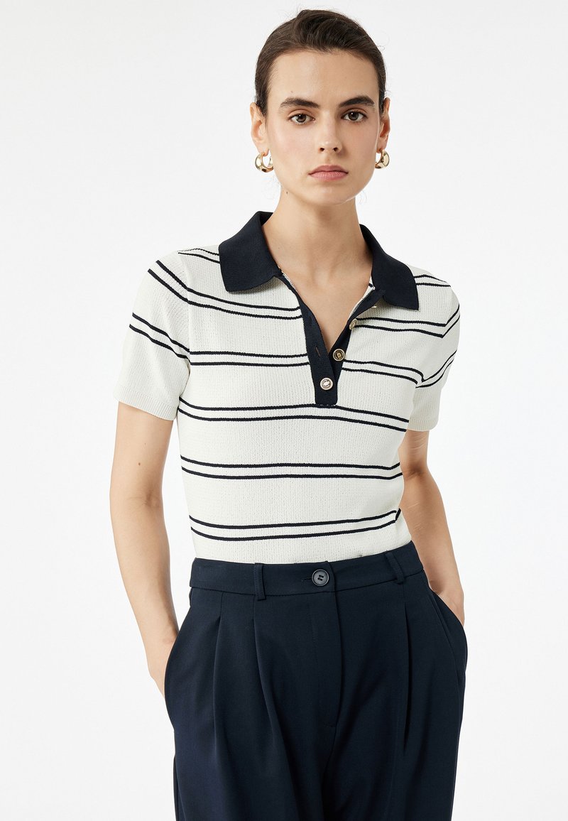 Hvid polo t-shirt med marineblå striber, korte ærmer og en kontrastfarvet krave. Har tre knapper ved halsudskæringen. Sammen med mørke bukser.