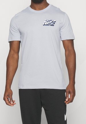 Homme à la peau mate portant un t-shirt Nike gris clair et un pantalon Nike noir, debout devant un fond blanc uni.